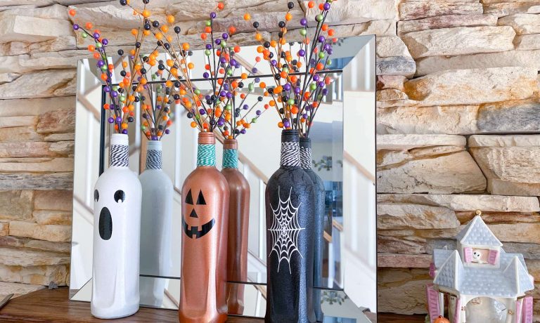 Halloween wine bottle craft on fireplace mantel with white ghost face bottle, orange pumpkin face bottle and black spider web bottle next to pastel Lenox haunted house
