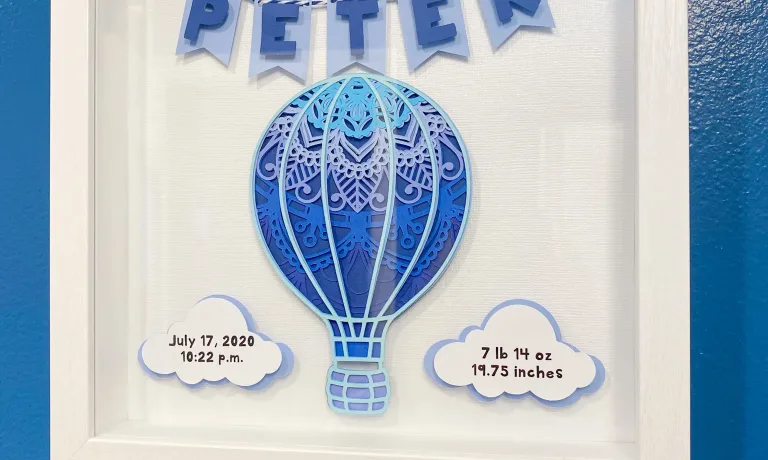 Layered hot air balloon mandala in shades of blue with date of birth and vital statistics on 3D clouds and a 3D pennant banner spelling out Peter strung with blue and white twine in a white shadowbox frame