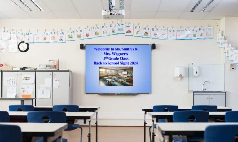 Classroom with desks in foreground facing whiteboard with blue slide that reads Welcome to Ms. Smith's and Mrs. Wagner's 5th Grade Class Back to School Night 2024, with alphabet above the board