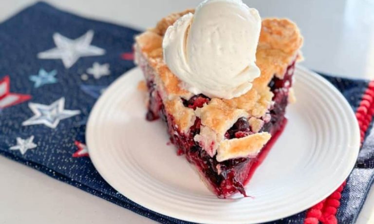 Slice of strawberry blueberry pie with scoop of vanilla ice cream on top on a white plate on top of a navy blue napkin with red, white and blue stars